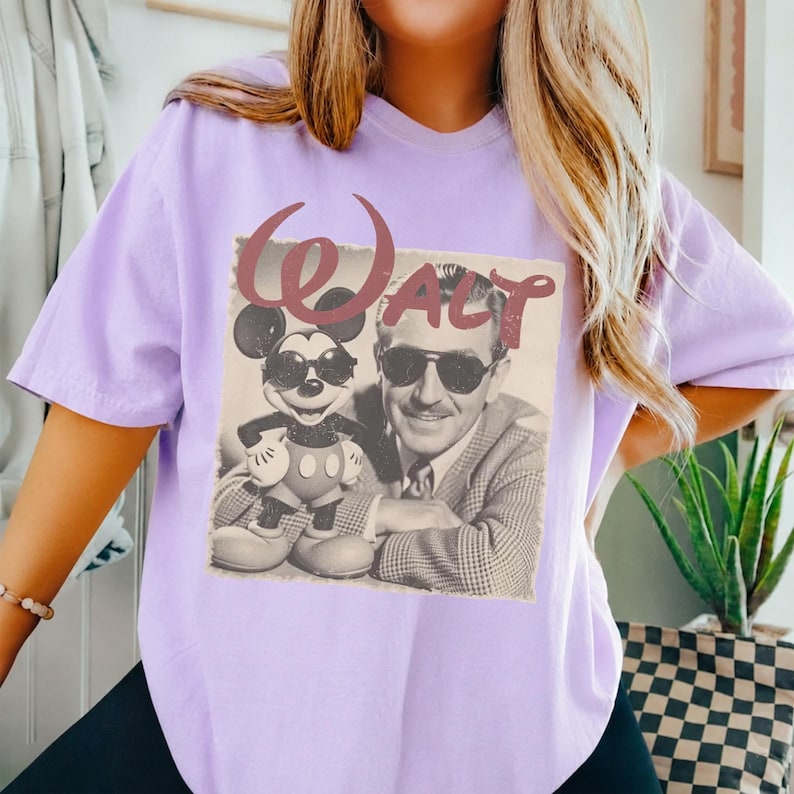 K&ouml;nnte beinhalten: Ein hellviolettes T-Shirt mit einem Vintage-Grafikdruck von Mickey Mouse und Walt Disney, die beide eine Sonnenbrille tragen. Der Text "WALT" ist in einer gro&szlig;en, stilisierten, rotbraunen Schrift &uuml;ber dem schwarz-wei&szlig;en Fotomotiv abgebildet.