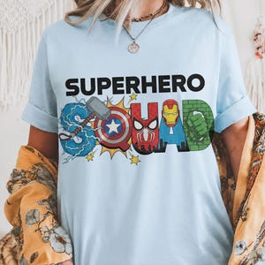 May include: Light blue t-shirt with the words "SUPERHERO SQUAD" in black block letters. The word "SQUAD" is designed with superhero logos and imagery, including Thor's hammer, Captain America's shield, and Spider-Man.