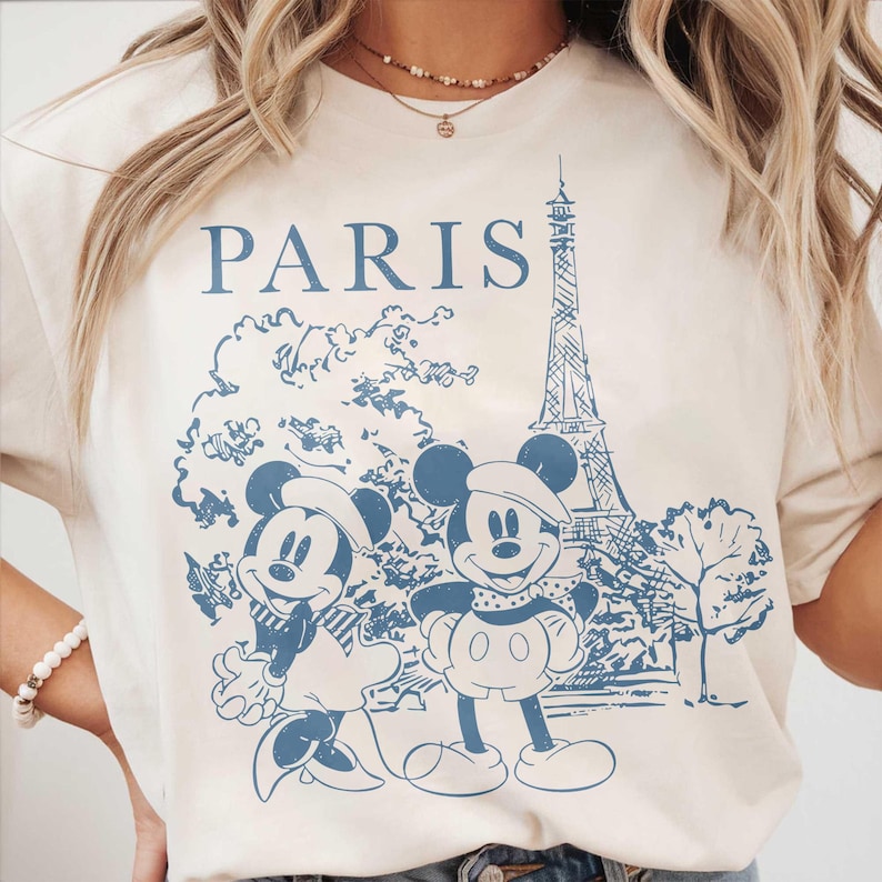 K&ouml;nnte beinhalten: Cremefarbenes T-Shirt mit einem blauen Aufdruck von Mickey und Minnie Mouse vor dem Eiffelturm. Das Wort "PARIS" ist &uuml;ber der Illustration aufgedruckt. Das Design hat eine Vintage-&Auml;sthetik mit Reisethema.