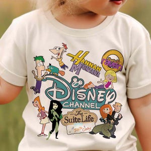 May include: Cream-coloured t-shirt featuring cartoon characters from Disney Channel shows. The shirt includes the Disney Channel logo and text for "Hannah Montana," "That's So Raven," and "The Suite Life of Zack & Cody."