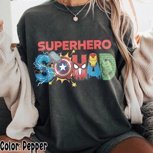 May include: Pepper-colored t-shirt with the words "SUPERHERO SQUAD" in red, blue, and green. The word "SQUAD" is designed with superhero logos and symbols. The shirt is a crew neck and has a relaxed fit.