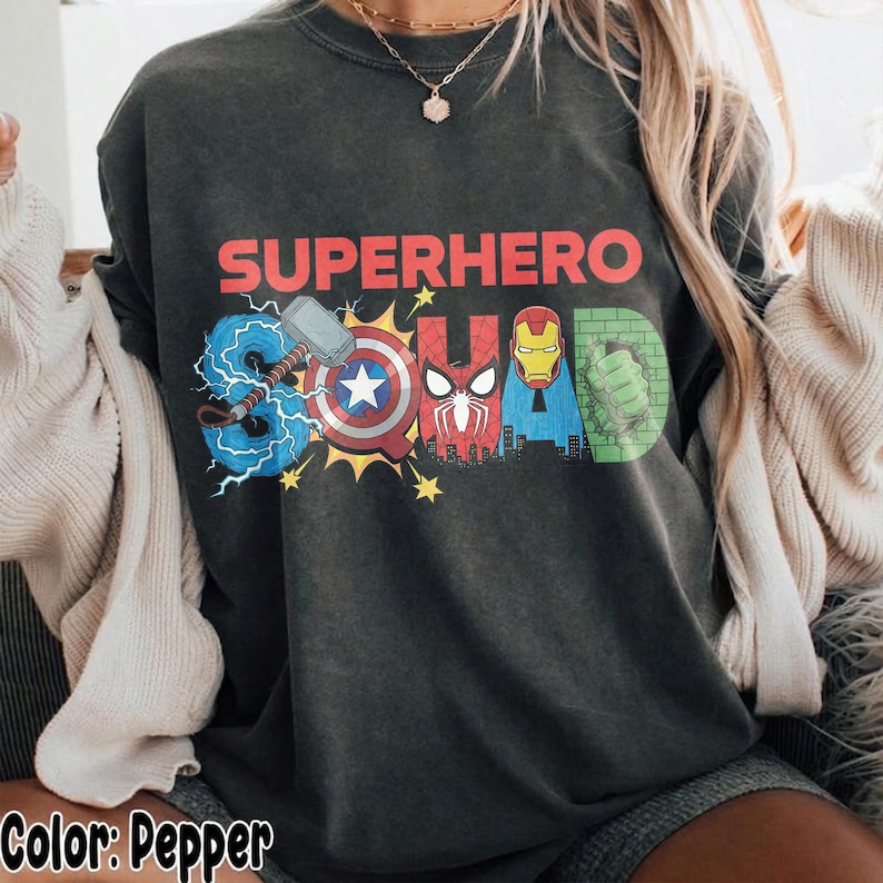 May include: Pepper-colored t-shirt with the words "SUPERHERO SQUAD" in red, blue, and green. The word "SQUAD" is designed with superhero logos and symbols. The shirt is a crew neck and has a relaxed fit.