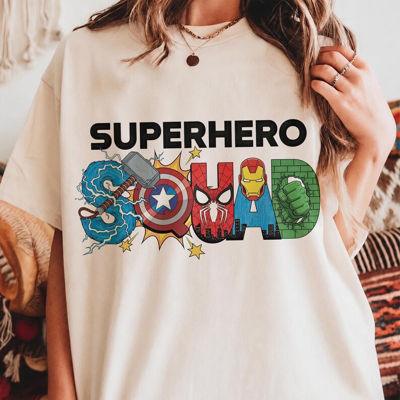 May include: Cream-colored t-shirt with the words "SUPERHERO SQUAD" in black block letters. The word "SQUAD" is designed with superhero-themed graphics, including Thor's hammer, Captain America's shield, and Iron Man's helmet.