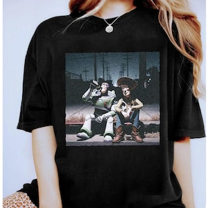 May include: Black t-shirt featuring a graphic of Buzz Lightyear and Woody from Toy Story, sitting on a curb. Buzz is drinking from a can, while Woody sits with his arms crossed. The image has a vintage, slightly faded look.