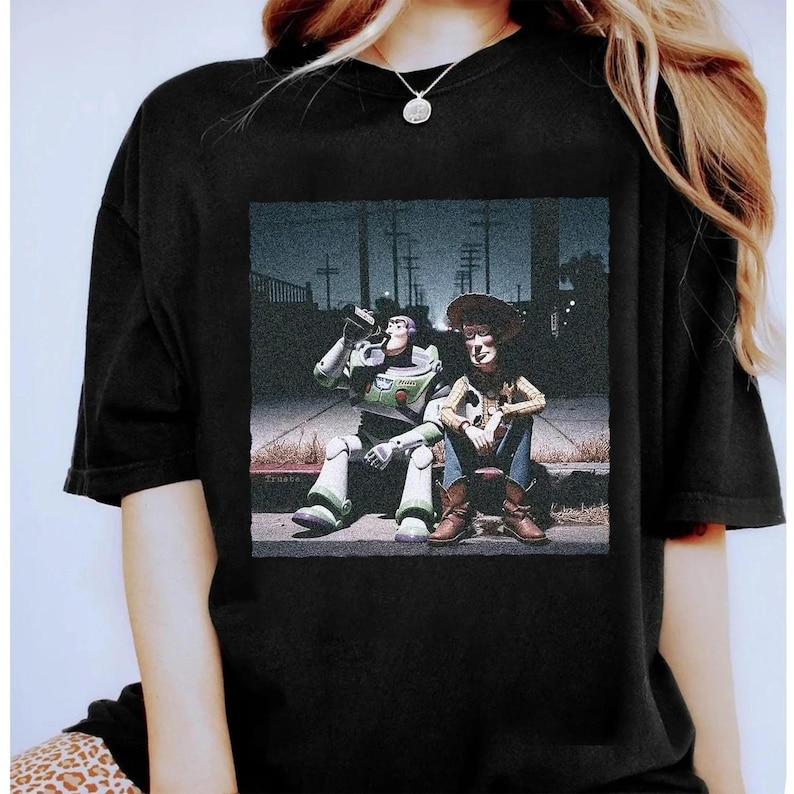 May include: Black t-shirt featuring a graphic of Buzz Lightyear and Woody from Toy Story, sitting on a curb. Buzz is drinking from a can, while Woody sits with his arms crossed. The image has a vintage, slightly faded look.