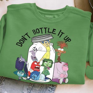 Peut inclure: Sweat-shirt vert avec l'inscription "DON'T BOTTLE IT UP". Le motif représente des personnages du film Vice-Versa dans un bocal. Le sweat-shirt est en matière douce et convient aux tenues décontractées.