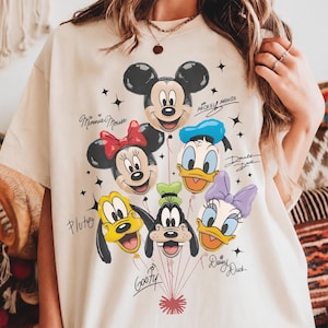 May include: A beige t-shirt featuring a graphic print of six Disney character heads designed as balloons, including Mickey Mouse, Minnie Mouse, Donald Duck, Pluto, Goofy, and Daisy Duck, with their names written in cursive script and small star accents.