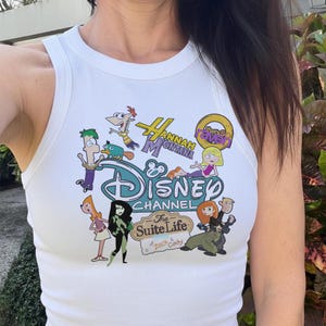 May include: White ribbed vest top with a colourful Disney Channel graphic. The graphic includes characters and logos from various Disney shows, such as Hannah Montana, Kim Possible, and The Suite Life of Zack & Cody.