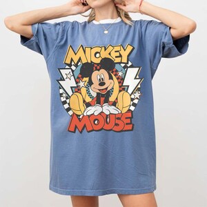 K&ouml;nnte beinhalten: Blaues T-Shirt mit einem Mickey Mouse-Motiv. Das Design zeigt die Worte "Mickey Mouse" in Gelb und Rot, mit Mickey in der Mitte. Das Shirt hat einen wei&szlig;en Kragen und kurze &Auml;rmel.