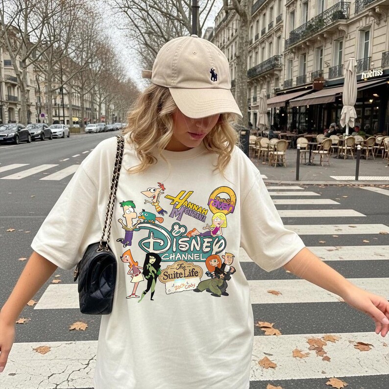 May include: A beige baseball cap with a dark blue logo and a white t-shirt featuring a Disney Channel graphic with cartoon characters from shows like Phineas and Ferb, Hannah Montana, and The Suite Life.