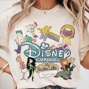 May include: A cream-coloured t-shirt with a Disney Channel graphic. The design features characters from "Hannah Montana", "That's So Raven", "The Suite Life of Zack & Cody", and "Kim Possible", along with the Disney Channel logo.
