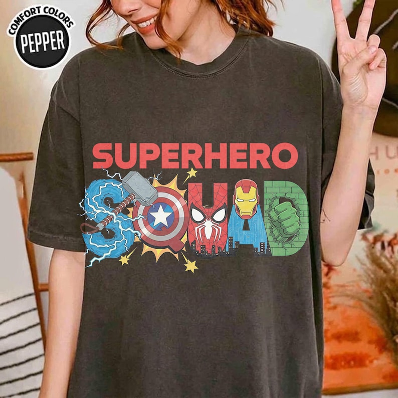 May include: Dark gray t-shirt with the words "SUPERHERO SQUAD" in red, blue, and green. The design incorporates superhero symbols, including a hammer, shield, and Iron Man's mask. The shirt has a vintage look.