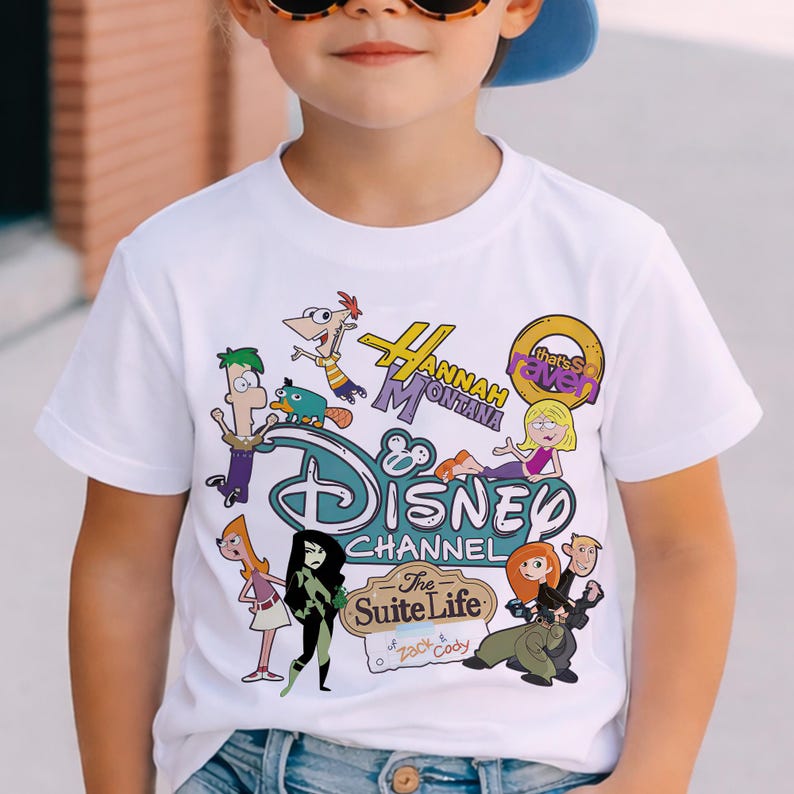 May include: White t-shirt with a vibrant Disney Channel design, showcasing characters from Hannah Montana, Phineas and Ferb, and Kim Possible. The top has a crew neck and short sleeves, designed for children's wear.
