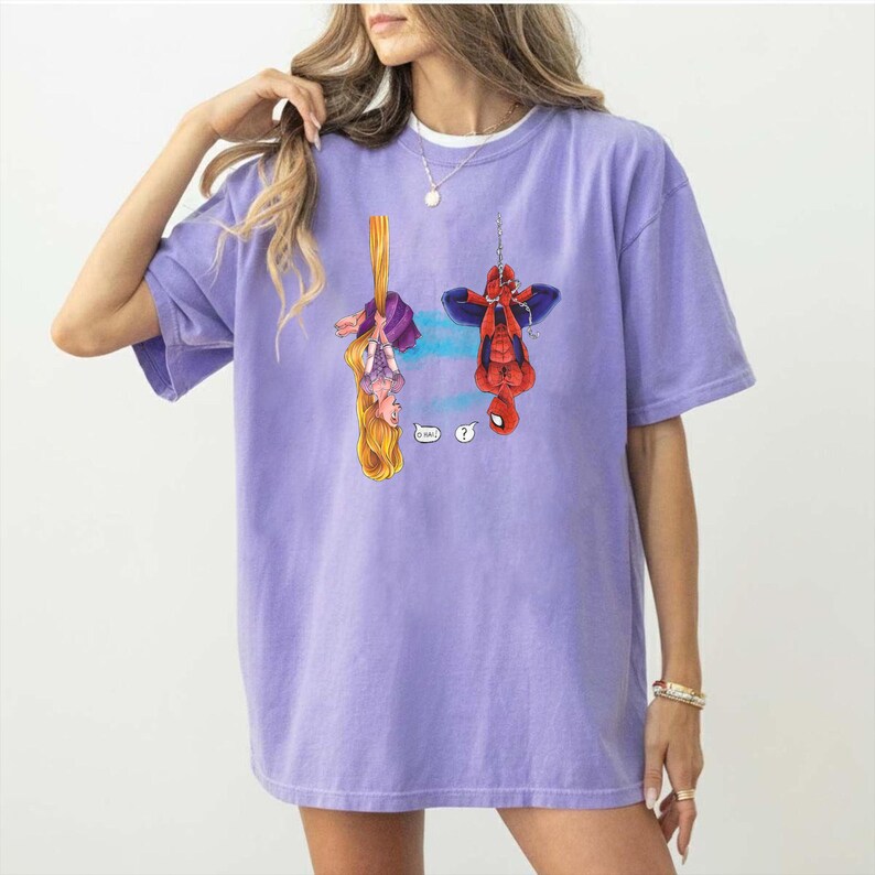 May include: A lilac-colored t-shirt featuring a cartoon illustration of Rapunzel and Spider-Man hanging upside down. Rapunzel is holding onto her long blonde hair, while Spider-Man is suspended by a web. Question mark speech bubbles are included.