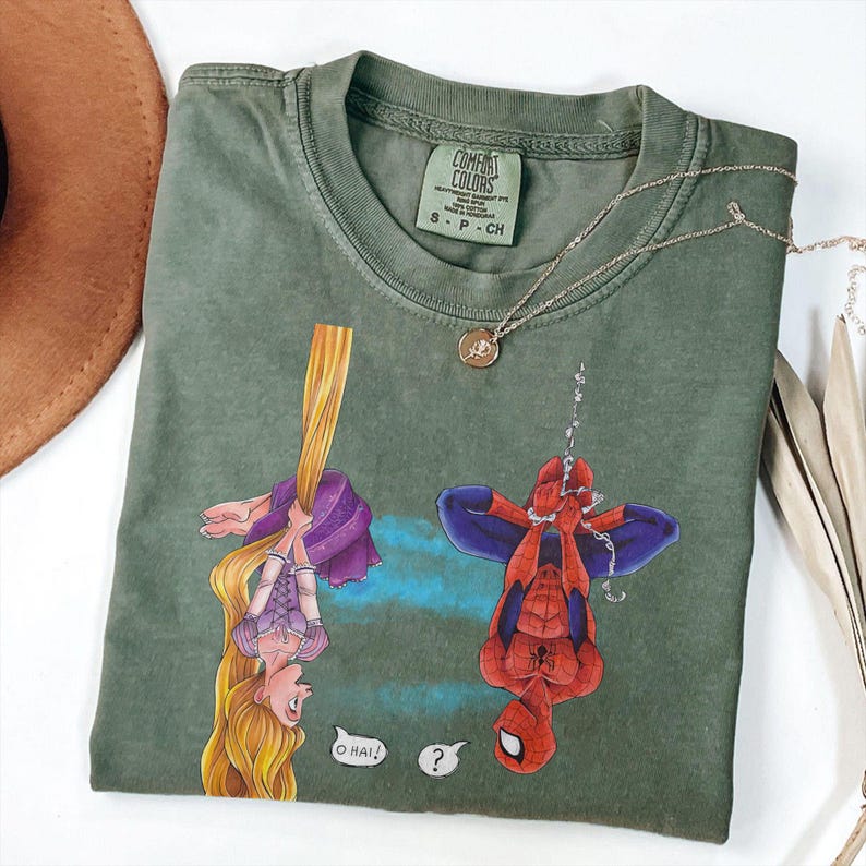 May include: Green Comfort Colors t-shirt featuring a cartoon of Rapunzel and Spider-Man. Rapunzel is hanging from her hair, and Spider-Man is upside down. The shirt has a small tag at the neckline and a gold necklace.