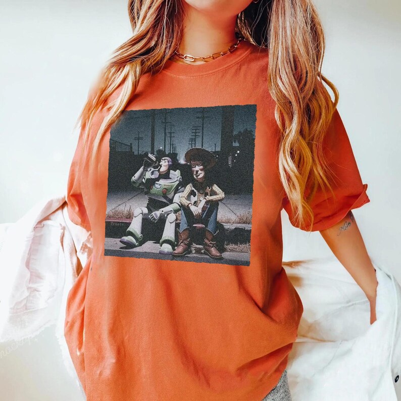May include: Burnt orange t-shirt featuring a graphic of Buzz Lightyear and Woody from Toy Story. The image shows the characters sitting on a curb. The shirt has short sleeves and a crew neck.