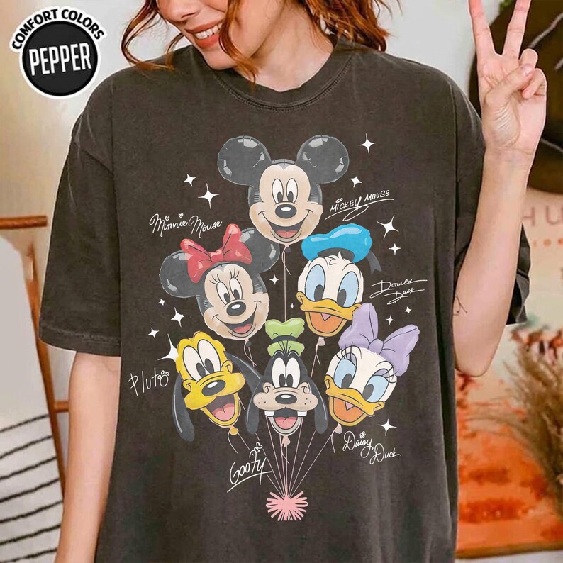 May include: A charcoal gray short-sleeve t-shirt featuring a graphic of Mickey Mouse, Minnie Mouse, Donald Duck, Pluto, Goofy, and Daisy Duck as balloons. The design includes white star accents and the names of each character in cursive script.