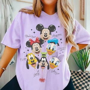 May include: A light purple t-shirt featuring a graphic print of Mickey Mouse, Minnie Mouse, Donald Duck, Pluto, Goofy, and Daisy Duck as balloons. Each character is labeled with their name in cursive script, surrounded by small black star accents.