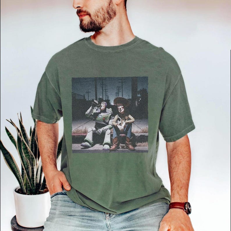May include: Green t-shirt featuring a graphic of two animated characters sitting on a curb. One character is a space ranger, and the other is a cowboy. The graphic is in a vintage style.