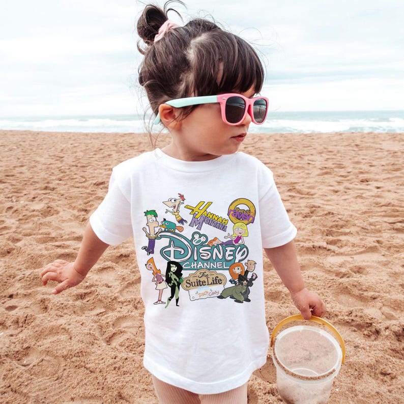 May include: A white t-shirt with a Disney Channel graphic featuring cartoon characters. The child is wearing pink sunglasses and holding a small bucket on a sandy beach. The graphic includes characters from various Disney shows.