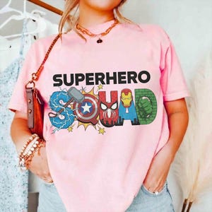 May include: Pink t-shirt with the words "SUPERHERO SQUAD" in black and colorful graphics. The "S" features Thor's hammer and lightning, the "U" has Captain America's shield, the "A" has Iron Man's helmet, and the "D" has the Hulk's fist.