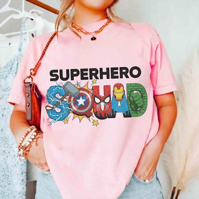 May include: Pink t-shirt with the words "SUPERHERO SQUAD" in black and colorful graphics. The "S" features Thor's hammer and lightning, the "U" has Captain America's shield, the "A" has Iron Man's helmet, and the "D" has the Hulk's fist.