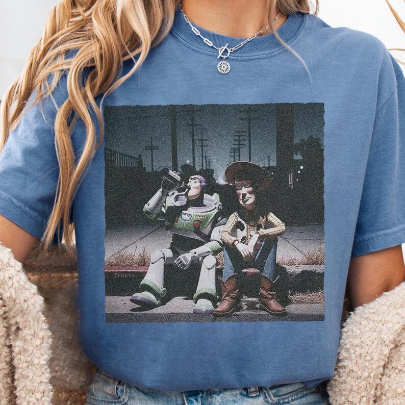 May include: Blue t-shirt featuring a graphic of Buzz Lightyear drinking from a bottle next to Woody, both sitting on a curb. The image has a vintage, slightly faded look, with a dark background and streetlights.