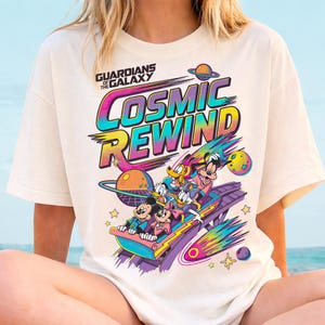 May include: Cream-colored t-shirt featuring a colorful graphic of Mickey Mouse, Donald Duck, and other characters on a roller coaster with the text "Cosmic Rewind" and "Guardians of the Galaxy".