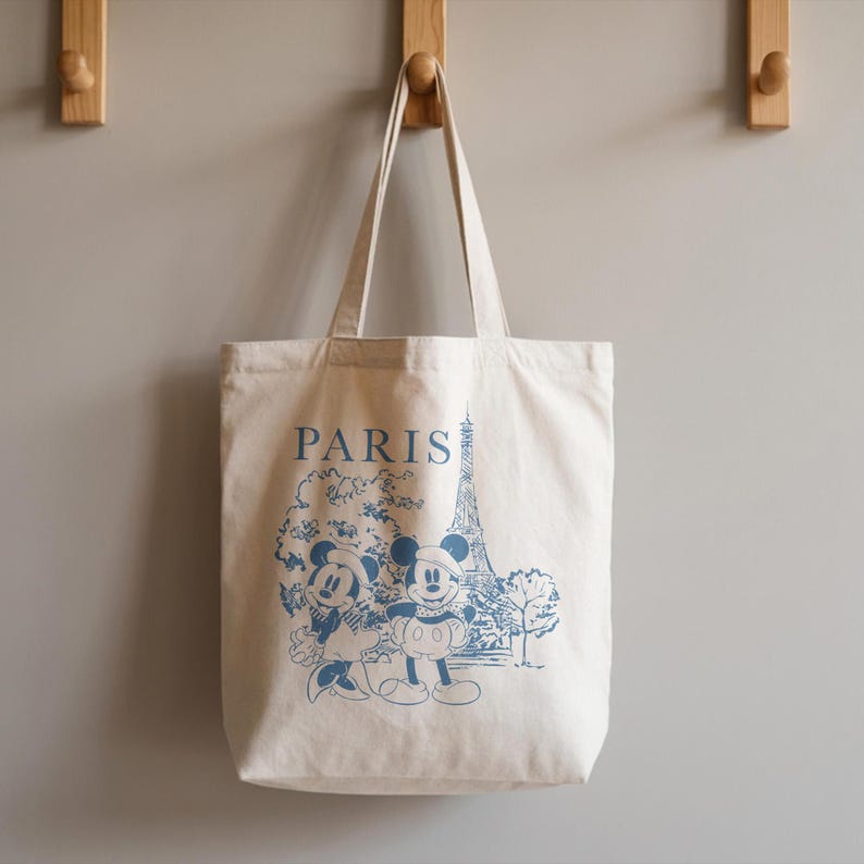K&ouml;nnte beinhalten: Cremefarbene Tragetasche mit blauem Aufdruck von Mickey und Minnie Mouse in Paris, mit dem Eiffelturm. Das Wort "PARIS" ist &uuml;ber der Illustration aufgedruckt. Die Tasche hat zwei Henkel und h&auml;ngt an einer Wand.