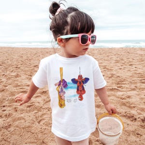 May include: White t-shirt featuring cartoon illustrations of Rapunzel and Spider-Man hanging upside down. Rapunzel is hanging from her long blonde hair, and Spider-Man is suspended by a web. The shirt is worn by a child on a sandy beach.