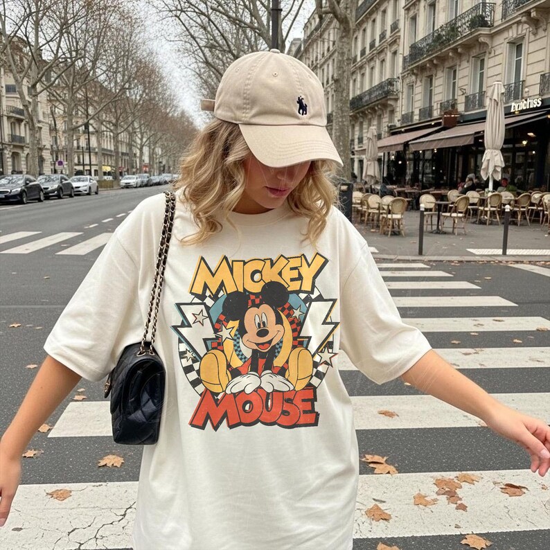 K&ouml;nnte beinhalten: Cremefarbenes T-Shirt mit einem farbenfrohen Mickey Mouse-Motiv und dem Text "MICKEY MOUSE". Eine beige Baseballkappe mit kleinem schwarzem Logo wird getragen. Eine schwarze Handtasche mit Kettenriemen ist ebenfalls zu sehen.