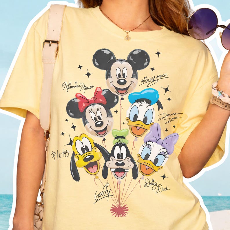 May include: A pale yellow short-sleeve t-shirt featuring a graphic print of Mickey Mouse, Minnie Mouse, Donald Duck, Pluto, Goofy, and Daisy Duck as balloons. Each character is labeled with their name in black script text, surrounded by small stars.