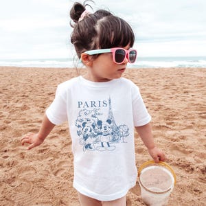 K&ouml;nnte beinhalten: Ein wei&szlig;es T-Shirt mit einem blauen Aufdruck von Mickey und Minnie Mouse vor dem Eiffelturm und dem Wort "PARIS". Das Kind tr&auml;gt eine rosa Sonnenbrille mit hellblauem Gestell.