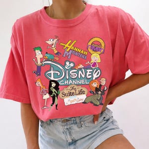 May include: A coral-coloured t-shirt with a Disney Channel graphic featuring cartoon characters from shows like Hannah Montana, Kim Possible, and Phineas and Ferb. The tee has a crew neck and short sleeves.