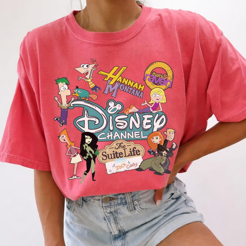 May include: A coral-coloured t-shirt with a Disney Channel graphic featuring cartoon characters from shows like Hannah Montana, Kim Possible, and Phineas and Ferb. The tee has a crew neck and short sleeves.
