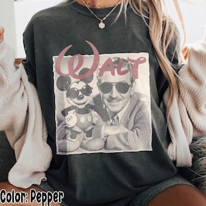 K&ouml;nnte beinhalten: Ein pfefferfarbenes T-Shirt mit Rundhalsausschnitt und einem Grafikdruck von Walt Disney und Mickey Mouse mit Sonnenbrillen. Der Schriftzug Walt ist in einer stilisierten rosa Schrift &uuml;ber den Figuren gedruckt. Kombiniert mit einer cremefarbenen Strickjacke.