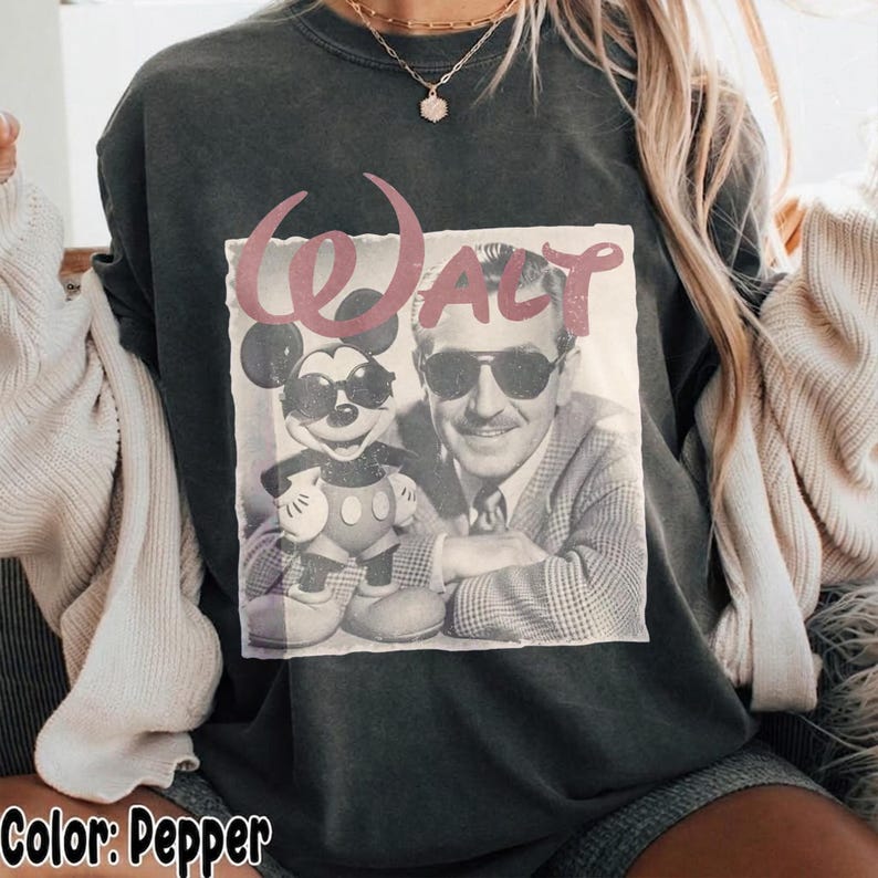 K&ouml;nnte beinhalten: Ein pfefferfarbenes T-Shirt mit Rundhalsausschnitt und einem Grafikdruck von Walt Disney und Mickey Mouse mit Sonnenbrillen. Der Schriftzug Walt ist in einer stilisierten rosa Schrift &uuml;ber den Figuren gedruckt. Kombiniert mit einer cremefarbenen Strickjacke.
