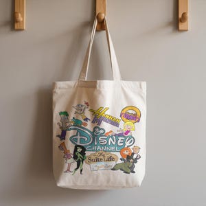 May include: A cream-coloured tote bag with a Disney Channel design. The bag features cartoon characters from shows such as Hannah Montana, Kim Possible, and Phineas and Ferb. The bag has two handles and is hanging on a wall, with the Disney Channel logo.