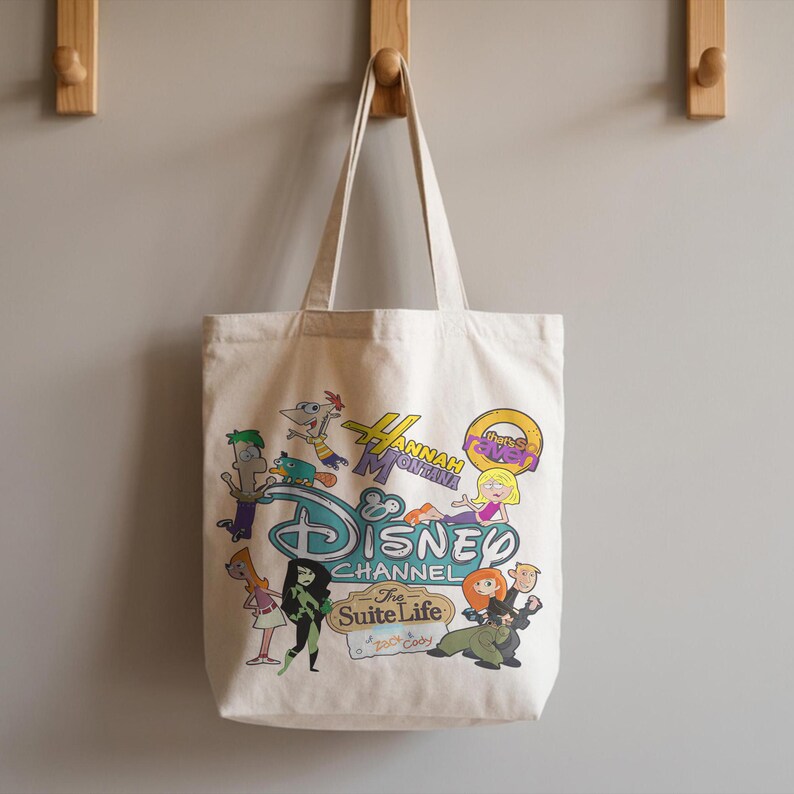 May include: A cream-coloured tote bag with a Disney Channel design. The bag features cartoon characters from shows such as Hannah Montana, Kim Possible, and Phineas and Ferb. The bag has two handles and is hanging on a wall, with the Disney Channel logo.