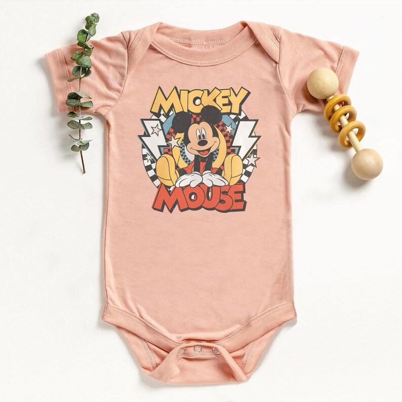 K&ouml;nnte beinhalten: Ein hellrosa Baby-Body mit einer Mickey Mouse-Grafik. Das Design enth&auml;lt die Worte "Mickey Mouse" in einer Retro-Schriftart. Eine Holzrassel mit gelben Ringen liegt neben dem Body.