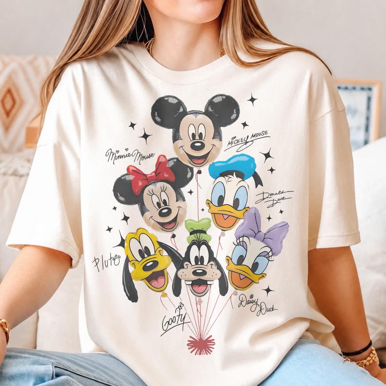 May include: A cream-colored short-sleeve t-shirt featuring a graphic of six Disney character balloons: Mickey Mouse, Minnie Mouse, Donald Duck, Pluto, Goofy, and Daisy Duck. Each character is labeled with their name in black cursive script.