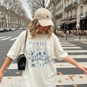 K&ouml;nnte beinhalten: Cremefarbenes T-Shirt mit blauem Aufdruck von Mickey und Minnie Mouse vor dem Eiffelturm, dar&uuml;ber das Wort "PARIS". Eine beige Baseballkappe mit dunkelblauem Logo wird getragen.