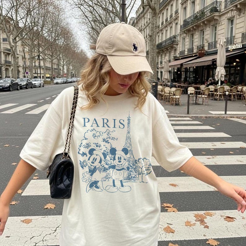 K&ouml;nnte beinhalten: Cremefarbenes T-Shirt mit blauem Aufdruck von Mickey und Minnie Mouse vor dem Eiffelturm, dar&uuml;ber das Wort "PARIS". Eine beige Baseballkappe mit dunkelblauem Logo wird getragen.