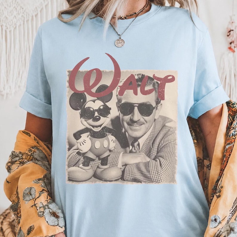 K&ouml;nnte beinhalten: Ein hellblaues T-Shirt mit einem grafischen Druck im Vintage-Stil, der Walt Disney und Mickey Mouse mit Sonnenbrillen zeigt. Das Wort "WALT" ist in einer gro&szlig;en, stilisierten roten Schrift &uuml;ber dem schwarz-wei&szlig;en Fotomotiv abgedruckt.