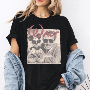 K&ouml;nnte beinhalten: Ein schwarzes kurz&auml;rmeliges T-Shirt mit einem Grafikdruck von Walt Disney und Mickey Mouse, die beide eine Sonnenbrille tragen. &Uuml;ber den Figuren ist das Wort WALT in einer stilisierten, abgenutzten rosa Schriftart aufgedruckt.