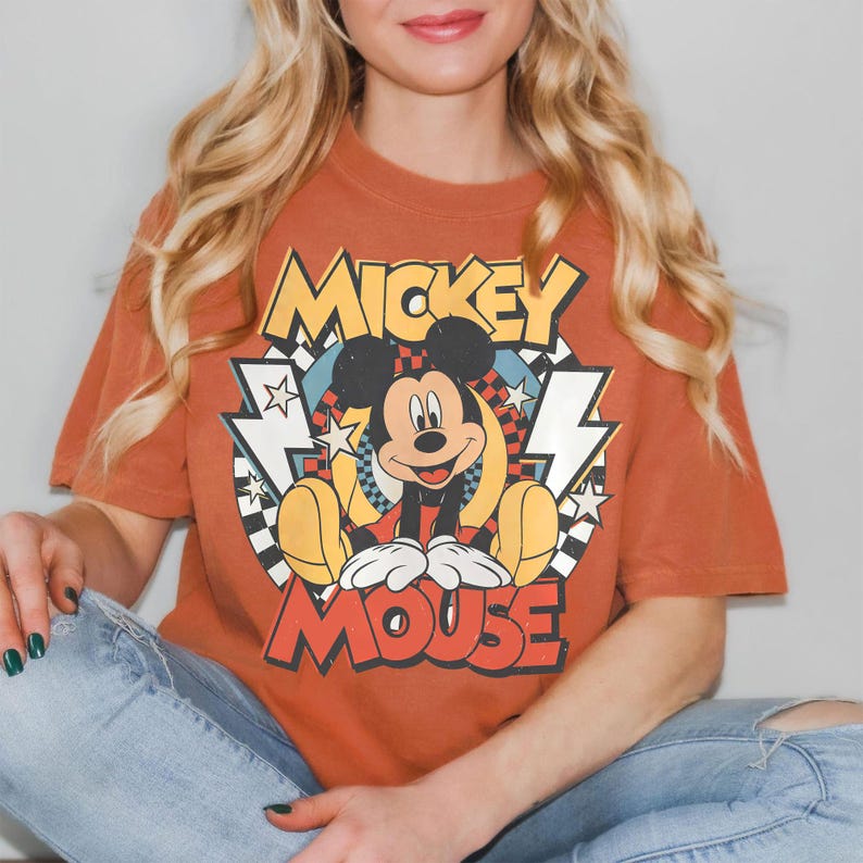 K&ouml;nnte beinhalten: Gebrannt orangefarbenes T-Shirt mit einem Vintage-Mickey-Mouse-Motiv. Das Design zeigt die Worte "Mickey Mouse" in Gelb und Rot, mit einem Cartoon-Bild von Mickey. Das T-Shirt wird mit einer blauen Jeans getragen.
