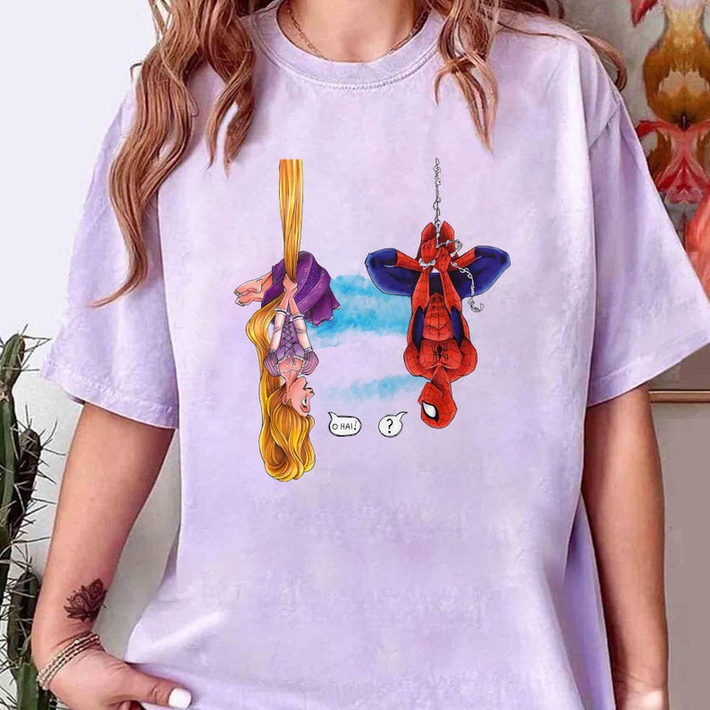 May include: A light purple t-shirt featuring a cartoon illustration of Rapunzel hanging from her hair and Spider-Man hanging upside down. Rapunzel has long blonde hair and is wearing a purple dress. Spider-Man is in his red and blue suit.
