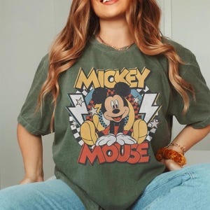 K&ouml;nnte beinhalten: Olivgr&uuml;nes T-Shirt mit einem Mickey Mouse-Motiv. Das Design zeigt die Worte "Mickey Mouse" in Gelb und Rot, zusammen mit einem Cartoon-Bild von Mickey. Das T-Shirt hat kurze &Auml;rmel und einen Rundhalsausschnitt.