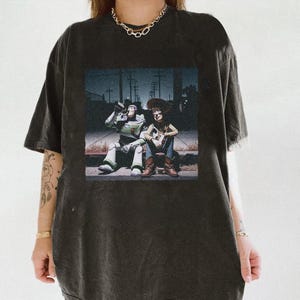 May include: Dark gray t-shirt featuring a graphic of Buzz Lightyear and Woody from Toy Story sitting together. The image is set against a nighttime street scene. The shirt has a distressed look.