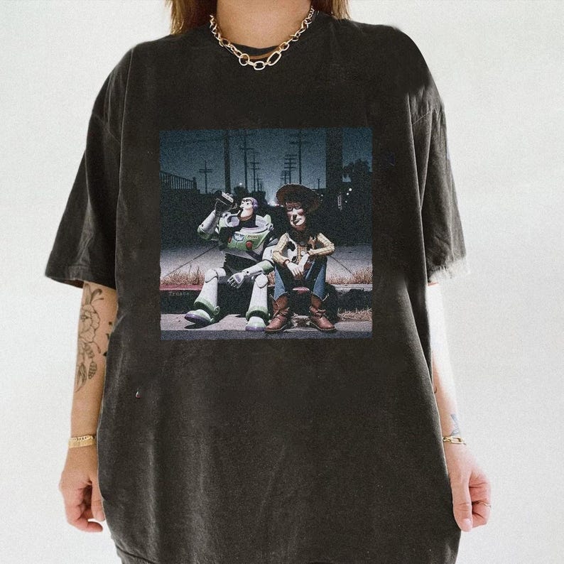 May include: Dark gray t-shirt featuring a graphic of Buzz Lightyear and Woody from Toy Story sitting together. The image is set against a nighttime street scene. The shirt has a distressed look.